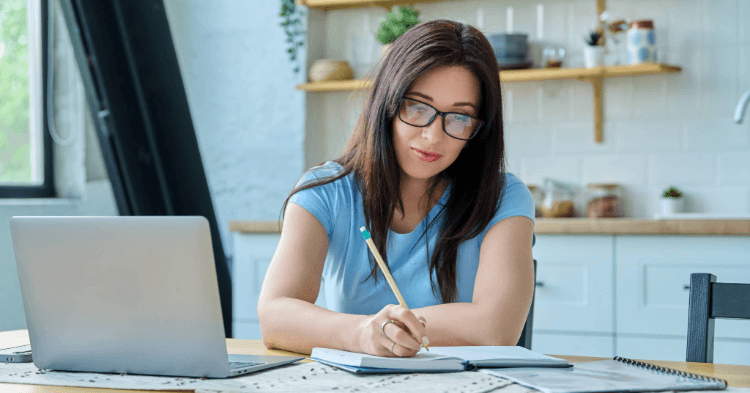 Woman sitting at a kitchen table with a laptop and note book writing