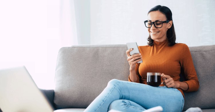 Woman wearing glasses and holding a cup of coffee sitting on the couch on her phone
