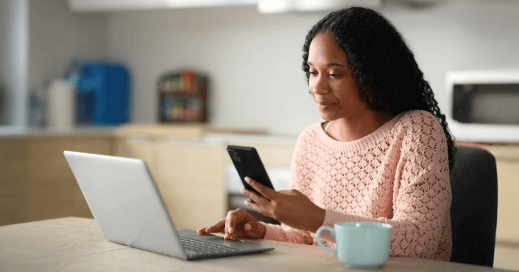 Woman at a kitchen table with laptop, phone and coffee mug