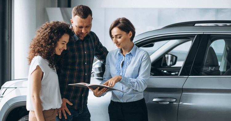 A couple and saleswoman look at a folder in front of a car.
