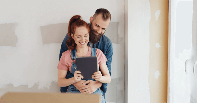 Young couple in the middle of home renovations hugging and looking at a tablet